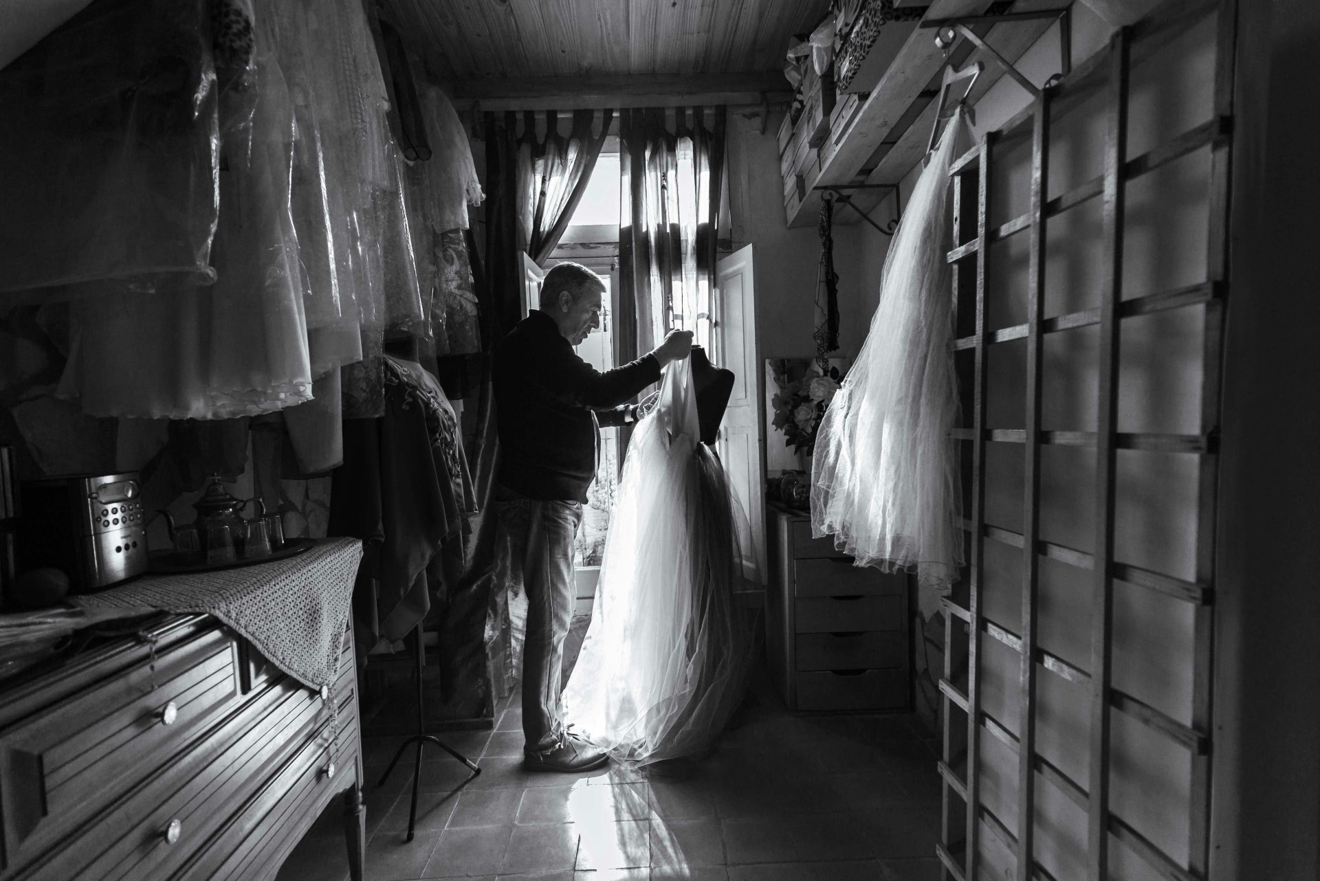 A tailor examines a wedding dress in a sunlit atelier filled with fabrics and dresses.