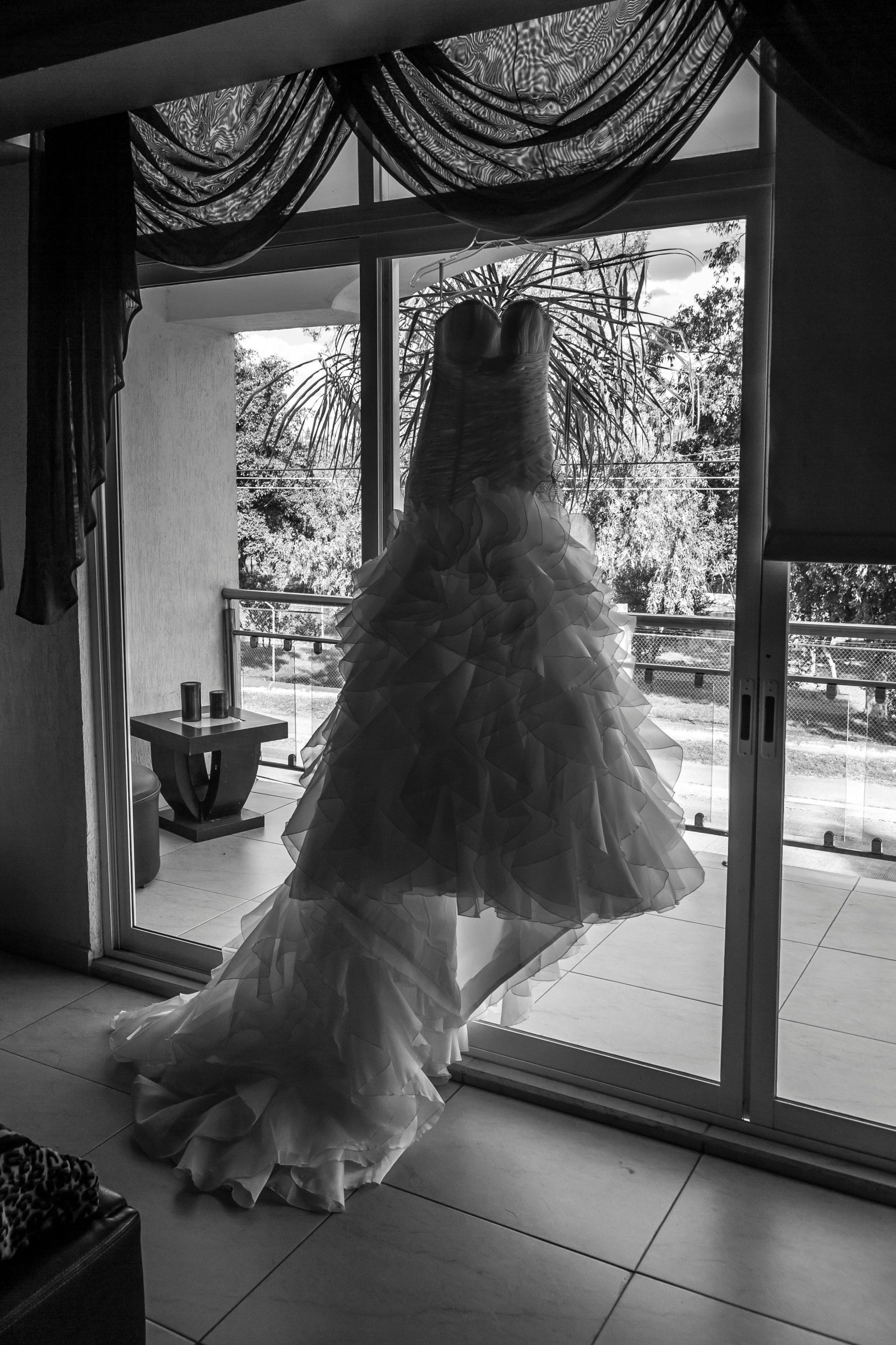 Black and white image of a wedding dress elegantly hung by a window, capturing a serene and romantic ambiance.